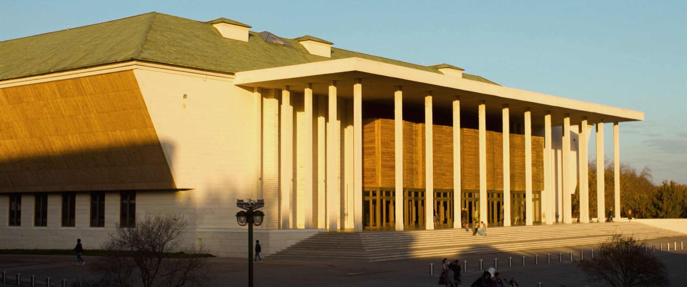 A modern civic public building at golden hour, people crossing the forecourt.