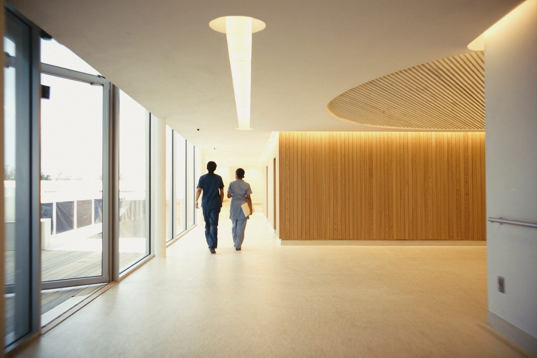 A bright hospital corridor in soft daylight with a clinician walking away from the camera.
