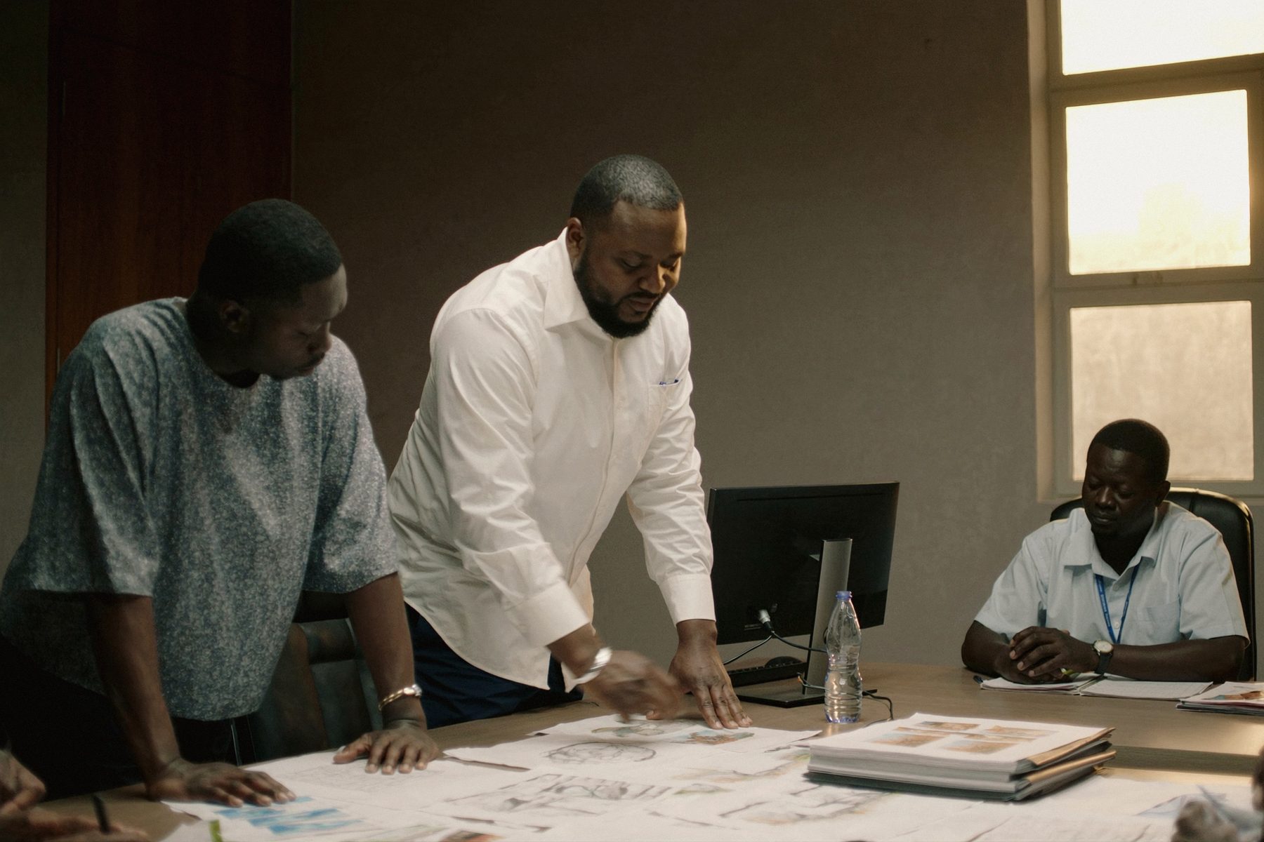 Three colleagues working over printed drawings and documents at a table in warm daylight.